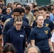 A large group of U.S. Navy personnel are participating in what appears to be a group walk or physical fitness event. The majority are wearing standard Navy physical training (PT) gear including navy blue t-shirts and hoodies with gold eagle insignia. A mix of active duty members and at least one retired CPO (Chief Petty Officer) are visible in the crowd.