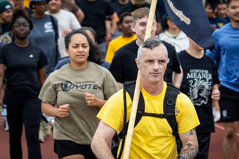 A large group of people are participating in what appears to be a military or organizational run/march event on a track. The man in the foreground is wearing a yellow shirt and black backpack while carrying a flag or guidon labeled 'The Lone Coyote.' Participants are wearing various shirts, including one referencing 'USS Pierre Coyote,' suggesting a Navy unit affiliation.