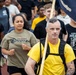 A large group of people are participating in what appears to be a military or organizational run/march event on a track. The man in the foreground is wearing a yellow shirt and black backpack while carrying a flag or guidon labeled 'The Lone Coyote.' Participants are wearing various shirts, including one referencing 'USS Pierre Coyote,' suggesting a Navy unit affiliation.