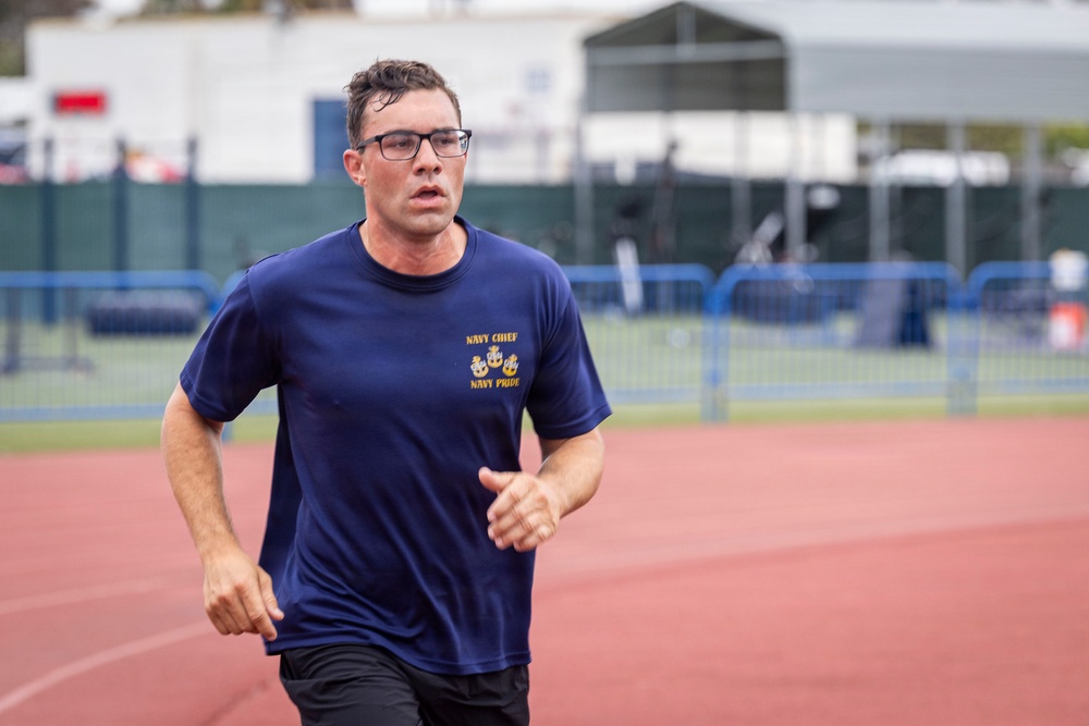 A man wearing glasses and a navy blue 'Navy Chief, Navy Pride' t-shirt is running on a red athletic track. He appears to be mid-stride with a focused expression, suggesting he is participating in a physical fitness test or training run. The background shows a blurred sports facility with blue fencing and white structures.