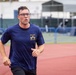 A man wearing glasses and a navy blue 'Navy Chief, Navy Pride' t-shirt is running on a red athletic track. He appears to be mid-stride with a focused expression, suggesting he is participating in a physical fitness test or training run. The background shows a blurred sports facility with blue fencing and white structures.