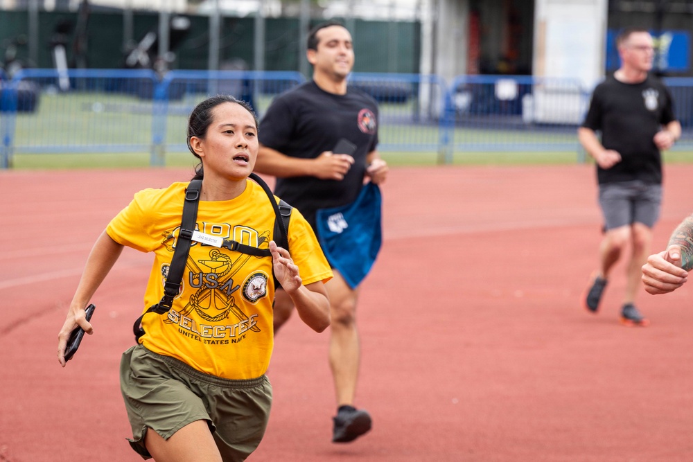 Three individuals are running on a red athletic track, with a woman in the foreground wearing a yellow 'United States Navy Selected' t-shirt and a backpack. She appears to be leading the group and is running with intensity. The setting appears to be a military or fitness assessment event based on the attire and context.