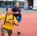 Three individuals are running on a red athletic track, with a woman in the foreground wearing a yellow 'United States Navy Selected' t-shirt and a backpack. She appears to be leading the group and is running with intensity. The setting appears to be a military or fitness assessment event based on the attire and context.