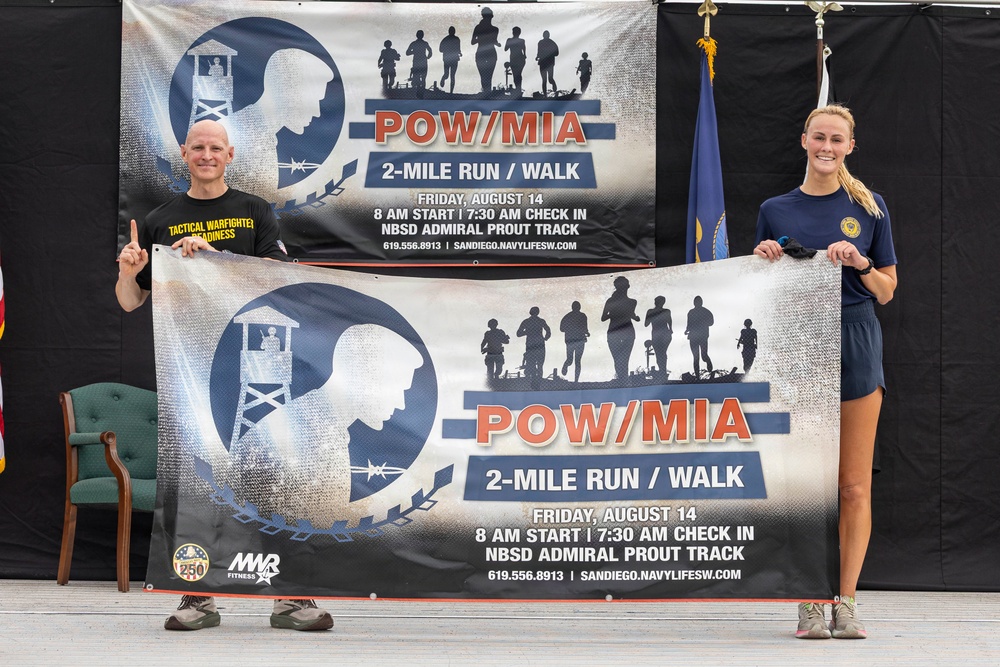 Two individuals are holding a promotional banner for a POW/MIA 2-Mile Run/Walk event scheduled for Friday, August 14, with an 8 AM start and 7:30 AM check-in at NBSD Admiral Prout Track in San Diego. A matching banner is displayed on the backdrop behind them. The event appears to be organized by MWR Fitness and is associated with Navy Life Southwest.