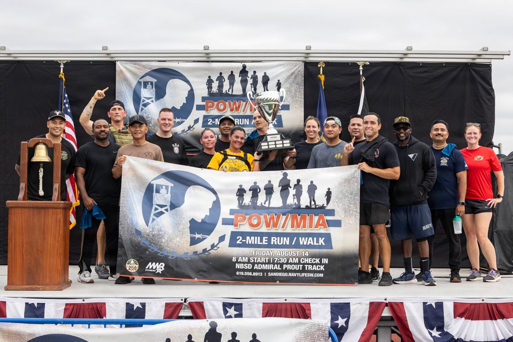 A group of approximately 15-20 people pose on a decorated stage at a POW/MIA 2-Mile Run/Walk event held at NBSD Admiral Prout Track in San Diego. Two individuals in the center are holding a large trophy cup, and the group is displaying a banner for the event. The event was scheduled for Friday, August 14, with an 8 AM start time.