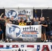 A group of approximately 15-20 people pose on a decorated stage at a POW/MIA 2-Mile Run/Walk event held at NBSD Admiral Prout Track in San Diego. Two individuals in the center are holding a large trophy cup, and the group is displaying a banner for the event. The event was scheduled for Friday, August 14, with an 8 AM start time.