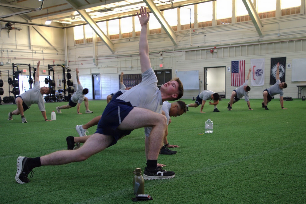 A group of individuals are performing a stretching or warm-up exercise in a large indoor athletic facility with artificial turf flooring. The foreground shows a young man performing a rotational lunge stretch with one arm raised overhead. An American flag and what appears to be a unit or organizational banner are visible on the far wall, suggesting a military or institutional training environment.