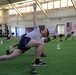 A group of individuals are performing a stretching or warm-up exercise in a large indoor athletic facility with artificial turf flooring. The foreground shows a young man performing a rotational lunge stretch with one arm raised overhead. An American flag and what appears to be a unit or organizational banner are visible on the far wall, suggesting a military or institutional training environment.