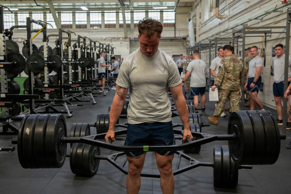 A muscular male athlete wearing a U.S. Air Force t-shirt performs a trap bar (hex bar) deadlift in a large military fitness facility. Multiple other personnel, some in military uniforms and some in PT gear, are visible in the background. The setting appears to be an official military physical fitness assessment or training event.
