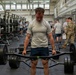 A muscular male athlete wearing a U.S. Air Force t-shirt performs a trap bar (hex bar) deadlift in a large military fitness facility. Multiple other personnel, some in military uniforms and some in PT gear, are visible in the background. The setting appears to be an official military physical fitness assessment or training event.