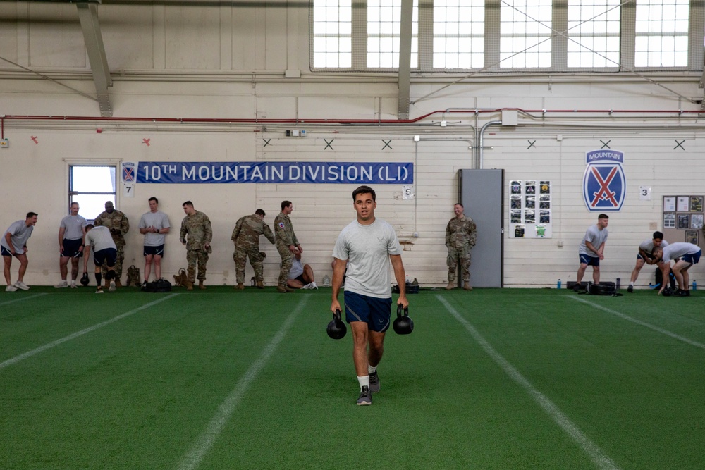 A soldier or service member carries two kettlebells across an indoor turf field facility belonging to the 10th Mountain Division (Light Infantry). Multiple personnel in both physical training attire and camouflage uniforms are visible in the background, suggesting a physical fitness assessment or training event. The facility is clearly marked with 10th Mountain Division banners and insignia.