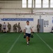 A soldier or service member carries two kettlebells across an indoor turf field facility belonging to the 10th Mountain Division (Light Infantry). Multiple personnel in both physical training attire and camouflage uniforms are visible in the background, suggesting a physical fitness assessment or training event. The facility is clearly marked with 10th Mountain Division banners and insignia.