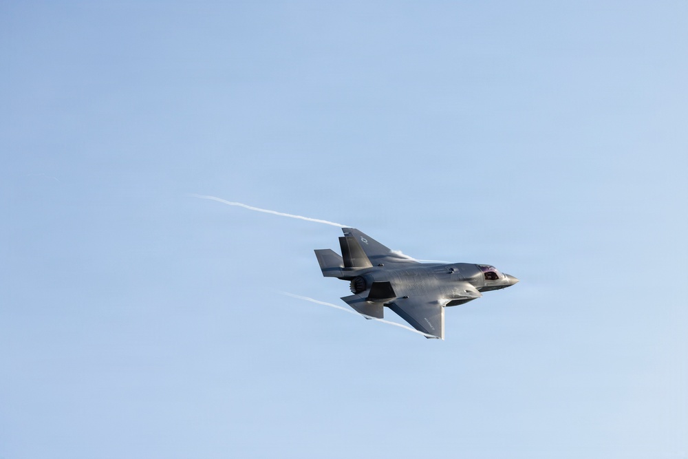 A single fifth-generation stealth fighter jet, consistent with an F-35 Lightning II, is captured in flight against a clear light-blue sky. The aircraft is banked slightly and traveling at high speed, producing visible condensation trails (wingtip vortices) streaming from its wings and tail section. The image is sharp and well-exposed, providing a clear view of the aircraft's distinctive delta-wing and stealth-optimized airframe design.