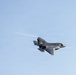 A single fifth-generation stealth fighter jet, consistent with an F-35 Lightning II, is captured in flight against a clear light-blue sky. The aircraft is banked slightly and traveling at high speed, producing visible condensation trails (wingtip vortices) streaming from its wings and tail section. The image is sharp and well-exposed, providing a clear view of the aircraft's distinctive delta-wing and stealth-optimized airframe design.
