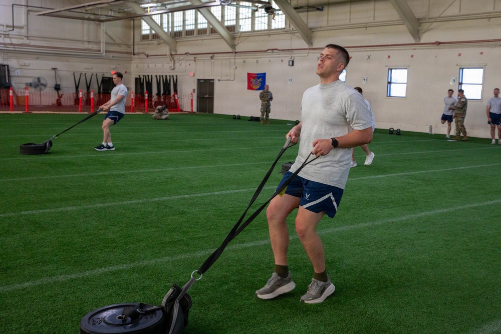 Military personnel are conducting a physical fitness training session inside a large indoor facility with artificial turf. The foreground shows a man in Air Force PT gear pulling a weighted sled using resistance straps, while others perform similar exercises in the background. A military unit flag or guidon is visible on the far wall, indicating an organized unit physical training event.