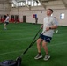Military personnel are conducting a physical fitness training session inside a large indoor facility with artificial turf. The foreground shows a man in Air Force PT gear pulling a weighted sled using resistance straps, while others perform similar exercises in the background. A military unit flag or guidon is visible on the far wall, indicating an organized unit physical training event.