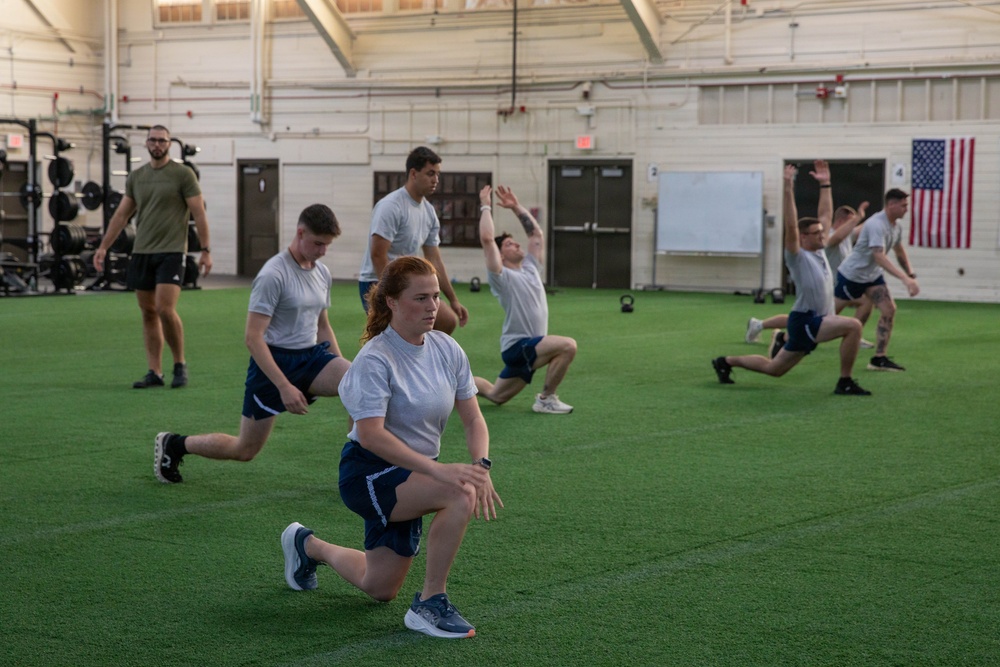 A group of individuals in matching white t-shirts and navy blue shorts perform lunge exercises on an indoor artificial turf facility. An instructor in an olive green shirt observes from the left side. The setting appears to be a military or institutional fitness facility, indicated by the American flag on the wall and the structured, uniform appearance of participants.