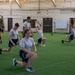A group of individuals in matching white t-shirts and navy blue shorts perform lunge exercises on an indoor artificial turf facility. An instructor in an olive green shirt observes from the left side. The setting appears to be a military or institutional fitness facility, indicated by the American flag on the wall and the structured, uniform appearance of participants.