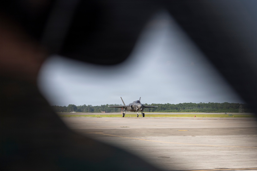 A military jet aircraft, consistent with an F-35 Lightning II, is taxiing on a runway or tarmac at a military airfield. The image is framed through what appears to be the gap in a person's arm or shoulder in the foreground, creating a natural vignette effect. Trees and overcast skies are visible in the background.