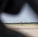 A military jet aircraft, consistent with an F-35 Lightning II, is taxiing on a runway or tarmac at a military airfield. The image is framed through what appears to be the gap in a person's arm or shoulder in the foreground, creating a natural vignette effect. Trees and overcast skies are visible in the background.