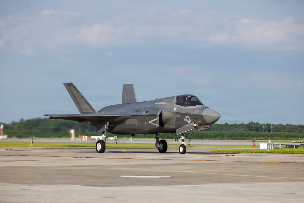 A U.S. military F-35 Lightning II stealth fighter jet is taxiing on an airfield tarmac. The aircraft is painted in a dark gray low-visibility scheme and displays U.S. roundel markings on the fuselage. The setting appears to be a military or joint-use airfield with green treeline visible in the background.