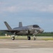 A U.S. military F-35 Lightning II stealth fighter jet is taxiing on an airfield tarmac. The aircraft is painted in a dark gray low-visibility scheme and displays U.S. roundel markings on the fuselage. The setting appears to be a military or joint-use airfield with green treeline visible in the background.