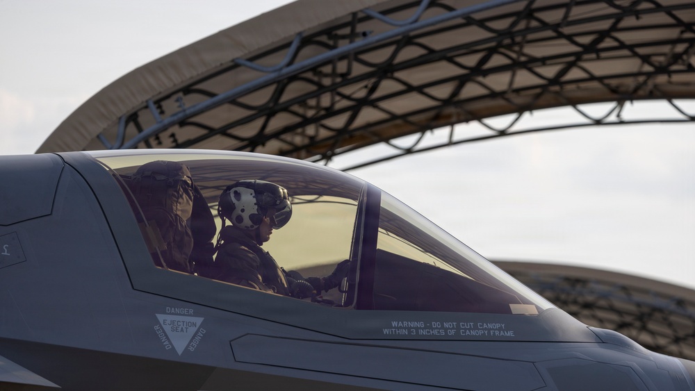 A military pilot is seated in the cockpit of what appears to be an F-35 Lightning II stealth fighter jet, visible through the closed canopy. The aircraft is positioned near a large curved hangar structure in the background. Safety markings are clearly visible on the canopy frame.