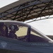 A military pilot is seated in the cockpit of what appears to be an F-35 Lightning II stealth fighter jet, visible through the closed canopy. The aircraft is positioned near a large curved hangar structure in the background. Safety markings are clearly visible on the canopy frame.