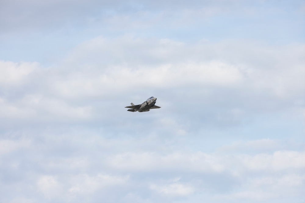 A single military jet aircraft is captured in flight against a partly cloudy sky. The aircraft appears to be a fifth-generation stealth fighter, consistent with an F-35 Lightning II design, flying at a moderate altitude. The image was taken from a ground-level perspective with the aircraft positioned near the center of the frame.