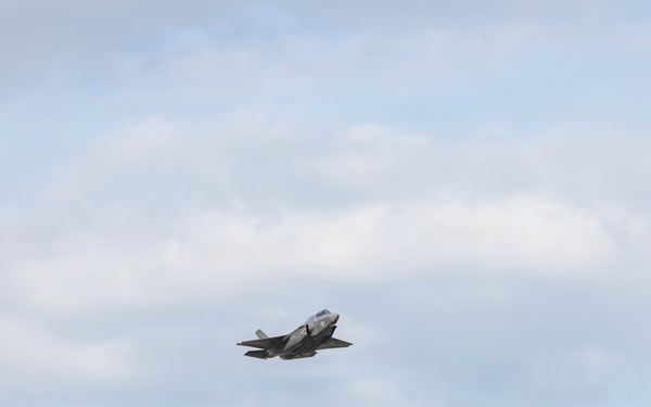 A single military jet aircraft is captured in flight against a partly cloudy sky. The aircraft appears to be a fifth-generation stealth fighter, consistent with an F-35 Lightning II design, flying at a moderate altitude. The image was taken from a ground-level perspective with the aircraft positioned near the center of the frame.