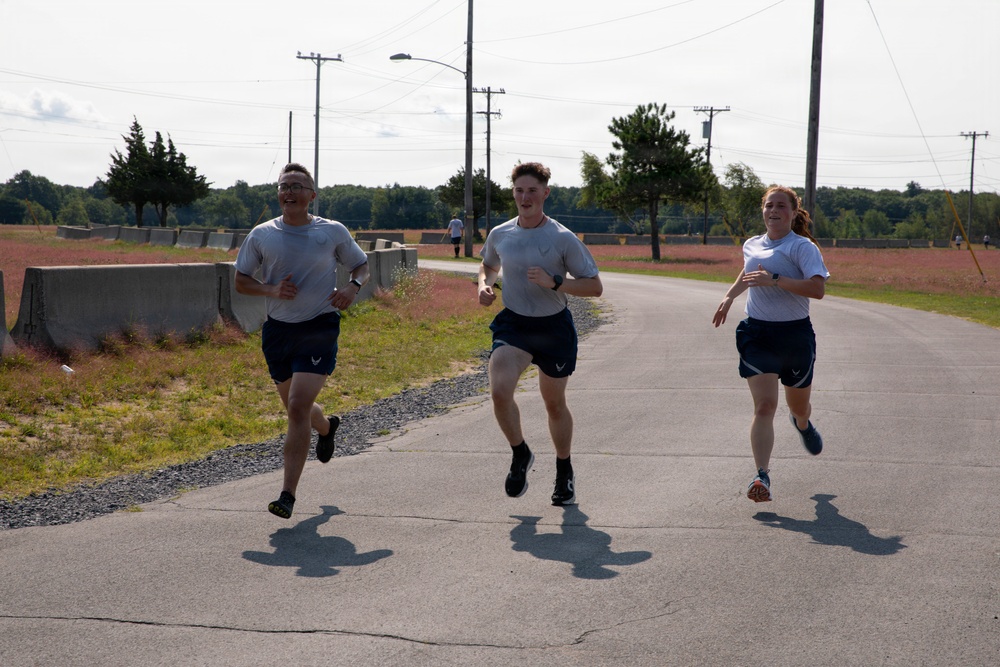 Three individuals in matching light gray t-shirts and dark navy shorts are running together on a paved road, appearing to be on a military base or training facility. All three are smiling and appear to be in good spirits during what looks like a physical fitness test or group run. The setting includes utility poles, concrete barriers, open fields, and tree lines in the background.
