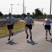 Three individuals in matching light gray t-shirts and dark navy shorts are running together on a paved road, appearing to be on a military base or training facility. All three are smiling and appear to be in good spirits during what looks like a physical fitness test or group run. The setting includes utility poles, concrete barriers, open fields, and tree lines in the background.