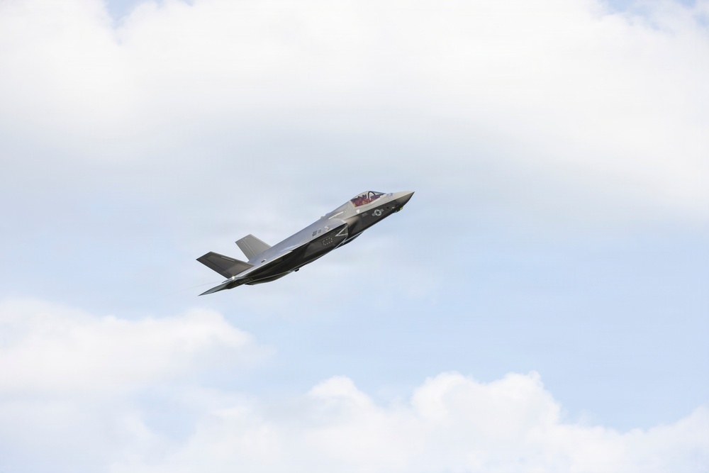 A single military stealth fighter jet is captured in flight against a partly cloudy sky. The aircraft appears to be an F-35 Lightning II, identifiable by its distinctive single-engine stealth design, delta wing configuration, and characteristic fuselage shape. The aircraft is banking slightly, suggesting a maneuver possibly during an airshow or training flight.