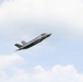 A single military stealth fighter jet is captured in flight against a partly cloudy sky. The aircraft appears to be an F-35 Lightning II, identifiable by its distinctive single-engine stealth design, delta wing configuration, and characteristic fuselage shape. The aircraft is banking slightly, suggesting a maneuver possibly during an airshow or training flight.