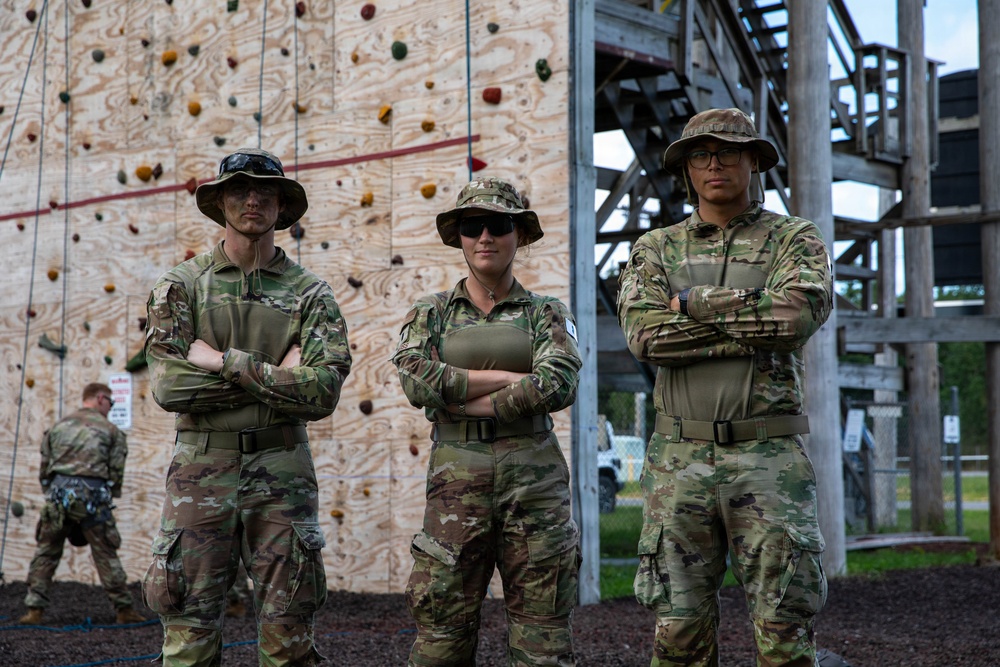 Three military personnel in matching OCP (Operational Camouflage Pattern) uniforms stand with arms crossed in front of a climbing wall at what appears to be a military training facility. All three wear boonie hats and sunglasses, presenting a unified, confident posture. A fourth service member is visible in the background near the climbing wall structure.