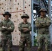 Three military personnel in matching OCP (Operational Camouflage Pattern) uniforms stand with arms crossed in front of a climbing wall at what appears to be a military training facility. All three wear boonie hats and sunglasses, presenting a unified, confident posture. A fourth service member is visible in the background near the climbing wall structure.