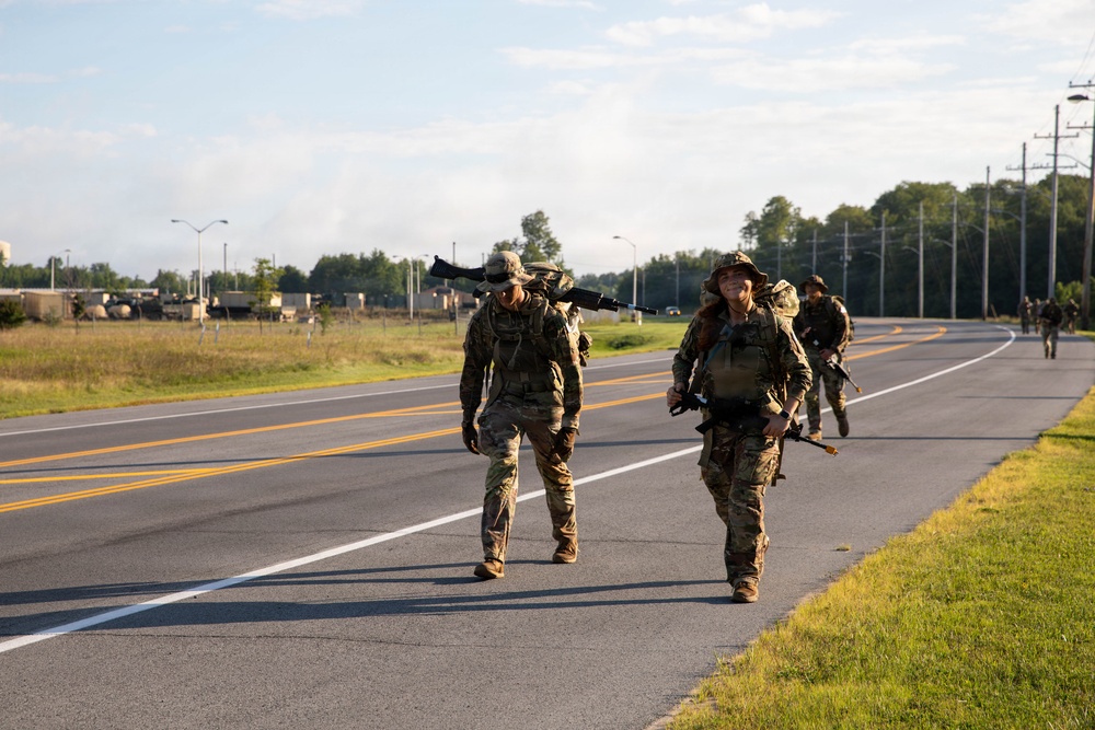 A group of military personnel in full combat gear are conducting a road march along a paved two-lane road, likely on a military installation. The soldiers are wearing camouflage uniforms and carrying rifles and rucksacks. Military vehicles are visible in the background, suggesting this is a training exercise or organized movement.