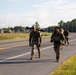 A group of military personnel in full combat gear are conducting a road march along a paved two-lane road, likely on a military installation. The soldiers are wearing camouflage uniforms and carrying rifles and rucksacks. Military vehicles are visible in the background, suggesting this is a training exercise or organized movement.