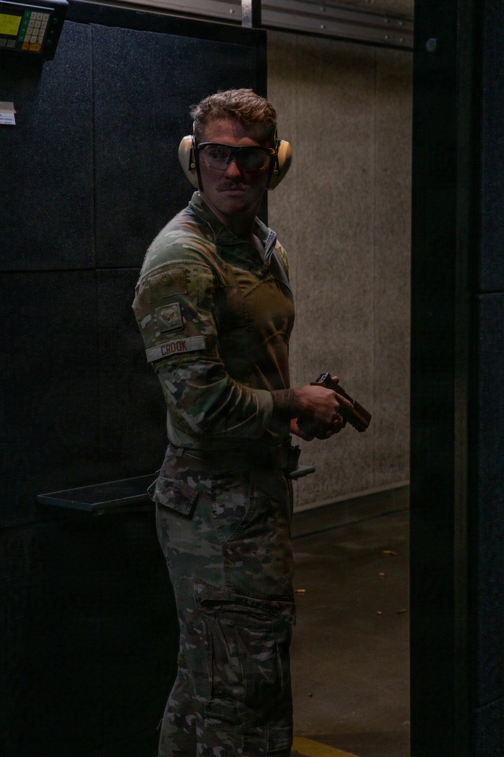 A military service member in MultiCam/OCP camouflage uniform is photographed at an indoor shooting range. The individual is wearing hearing protection (earmuffs) and safety glasses while holding a pistol in a low-ready or administrative position. A name tape reading 'CROOK' and a rank patch are visible on the left sleeve.