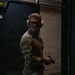 A military service member in MultiCam/OCP camouflage uniform is photographed at an indoor shooting range. The individual is wearing hearing protection (earmuffs) and safety glasses while holding a pistol in a low-ready or administrative position. A name tape reading 'CROOK' and a rank patch are visible on the left sleeve.