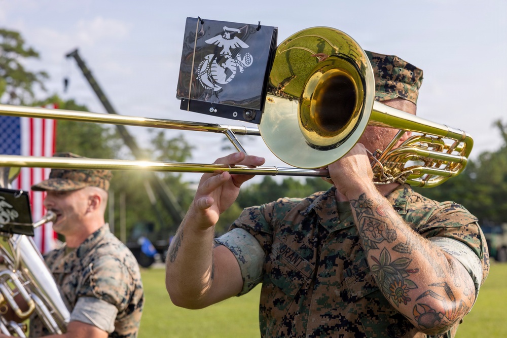 Two U.S. Marines in woodland MARPAT camouflage uniforms perform as part of a military band at an outdoor ceremony. The foreground Marine is playing a trombone equipped with a music stand holder displaying the Eagle, Globe, and Anchor emblem of the U.S. Marine Corps. An American flag is visible in the background, suggesting a formal military event or ceremony.