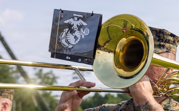 Two U.S. Marines in woodland MARPAT camouflage uniforms perform as part of a military band at an outdoor ceremony. The foreground Marine is playing a trombone equipped with a music stand holder displaying the Eagle, Globe, and Anchor emblem of the U.S. Marine Corps. An American flag is visible in the background, suggesting a formal military event or ceremony.