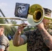 Two U.S. Marines in woodland MARPAT camouflage uniforms perform as part of a military band at an outdoor ceremony. The foreground Marine is playing a trombone equipped with a music stand holder displaying the Eagle, Globe, and Anchor emblem of the U.S. Marine Corps. An American flag is visible in the background, suggesting a formal military event or ceremony.
