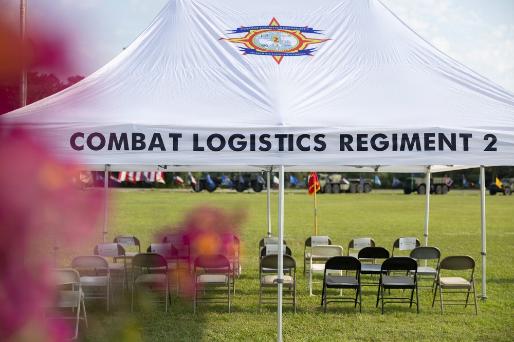 A white event tent bearing the insignia and name 'Combat Logistics Regiment 2' of the 2nd Marine Logistics Group is set up on a grassy field. Folding chairs are arranged in groups beneath the tent, suggesting preparation for a military ceremony or official event. In the background, flags, military vehicles, and additional event infrastructure are visible on the field.