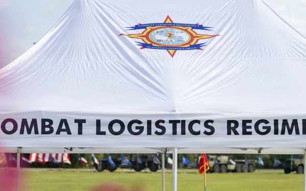 A white event tent bearing the insignia and name 'Combat Logistics Regiment 2' of the 2nd Marine Logistics Group is set up on a grassy field. Folding chairs are arranged in groups beneath the tent, suggesting preparation for a military ceremony or official event. In the background, flags, military vehicles, and additional event infrastructure are visible on the field.