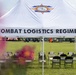 A white event tent bearing the insignia and name 'Combat Logistics Regiment 2' of the 2nd Marine Logistics Group is set up on a grassy field. Folding chairs are arranged in groups beneath the tent, suggesting preparation for a military ceremony or official event. In the background, flags, military vehicles, and additional event infrastructure are visible on the field.