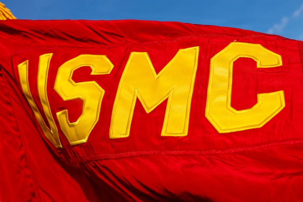 A close-up photograph of a red USMC (United States Marine Corps) flag flying against a blue sky. The flag features large yellow appliquéd letters 'USMC' sewn onto the scarlet red fabric. The flag appears to be in motion, showing visible ripples and folds in the material.