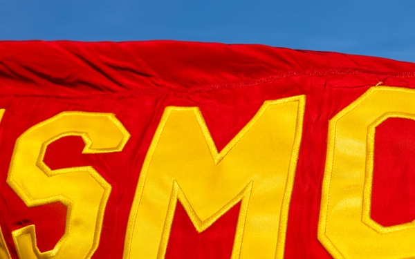 A close-up photograph of a red USMC (United States Marine Corps) flag flying against a blue sky. The flag features large yellow appliquéd letters 'USMC' sewn onto the scarlet red fabric. The flag appears to be in motion, showing visible ripples and folds in the material.