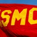 A close-up photograph of a red USMC (United States Marine Corps) flag flying against a blue sky. The flag features large yellow appliquéd letters 'USMC' sewn onto the scarlet red fabric. The flag appears to be in motion, showing visible ripples and folds in the material.