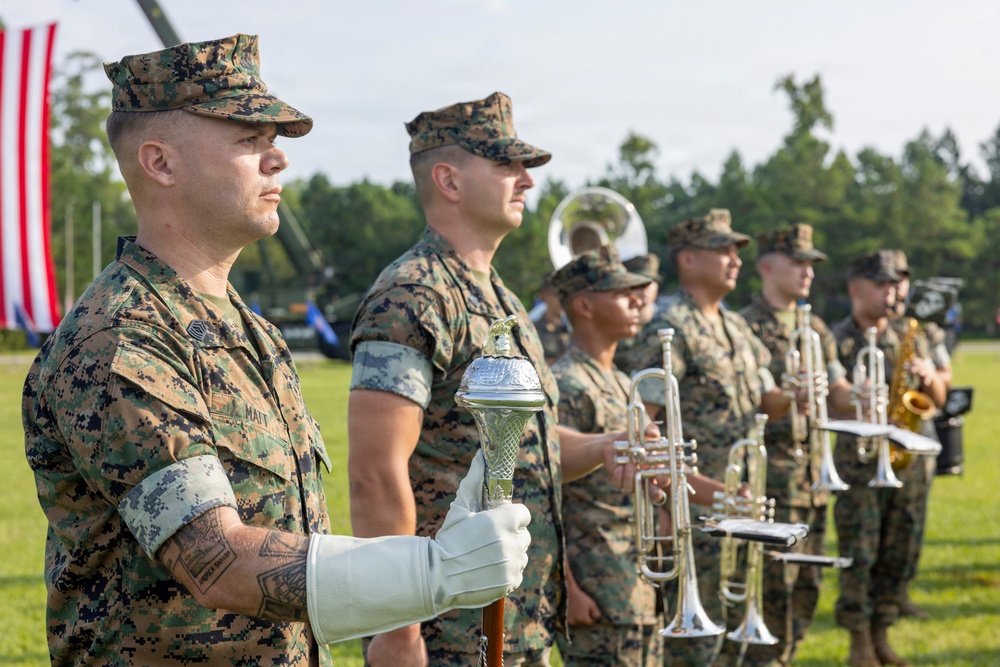 A group of U.S. Marines in digital camouflage (MARPAT) uniforms are standing at attention during a formal military ceremony on a grass field. The foreground Marine wears white ceremonial gloves and holds a mace or ceremonial staff, while behind him several Marines hold brass instruments including trumpets and a saxophone. An American flag is partially visible in the background along with trees and military equipment.